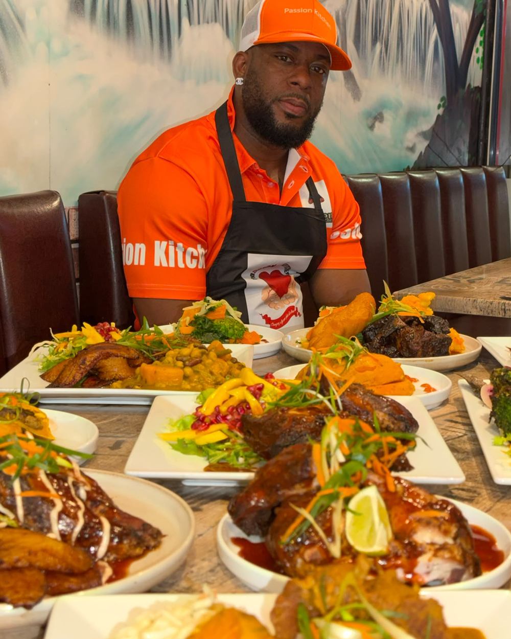 Chef Tyrell in the restaurant, in a Passion Kitchen shirt and apron, behind a long table of plated Jamaican dishes.