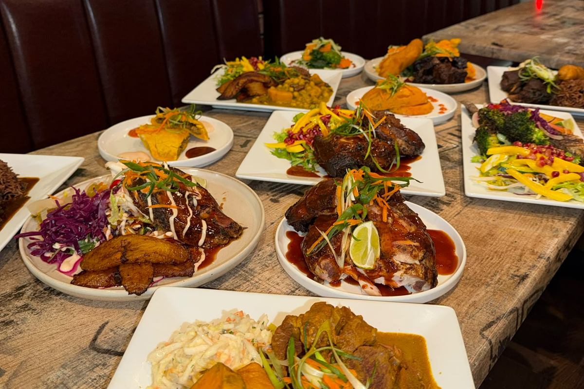A dozen plated dishes along a wooden table — jerk chicken with lime, curry goat with rice, fried plantain, coleslaw and mango salad.