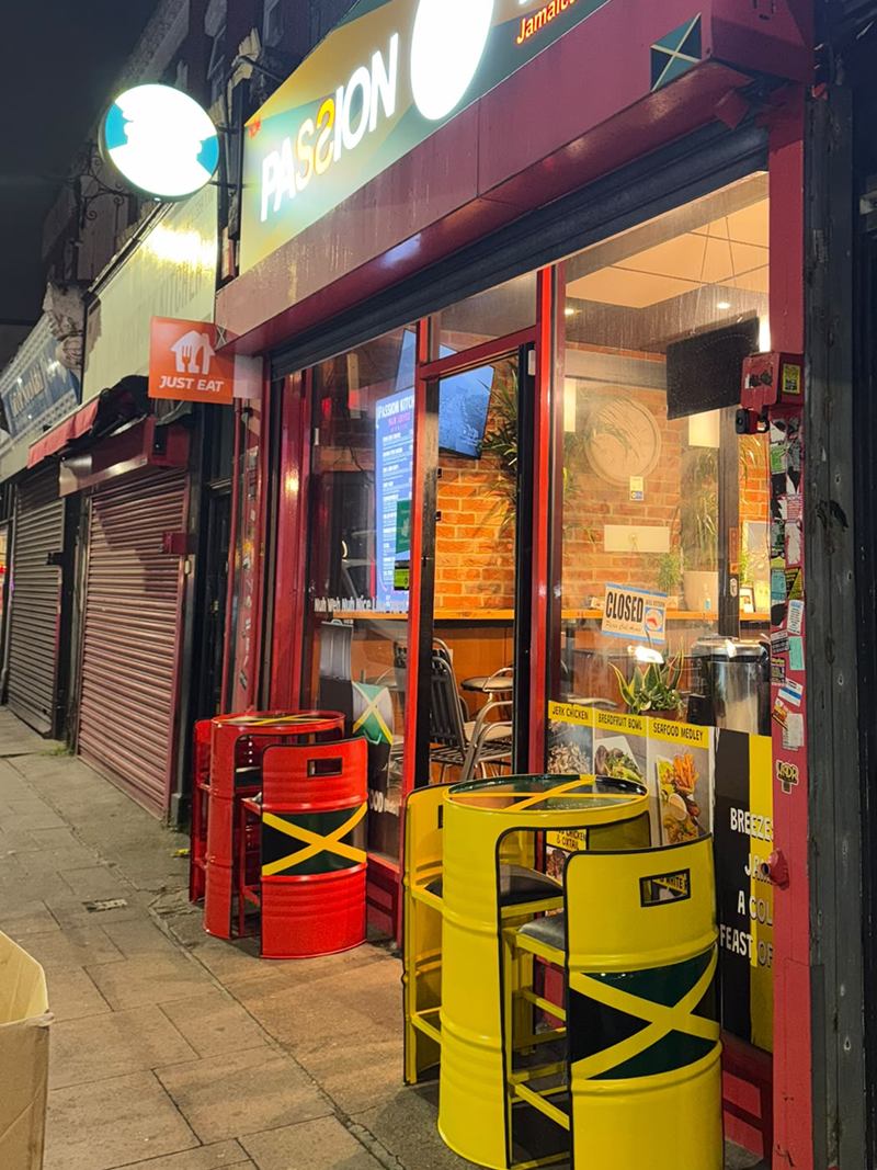 The Passion Kitchen shopfront at night, sign lit, with red and yellow Jamaican-flag barrel tables on the pavement outside.